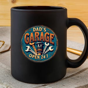 Może przedstawiać: Czarny ceramiczny kubek do kawy z grafiką vintage'owego znaku z napisem "Garage Taty Otwarty 24/7" z narzędziami i częścią samochodową w środku.