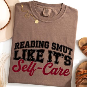May include: A brown t-shirt with the text "READING SMUT LIKE IT'S Self-Care" in black and burgundy lettering. A gold necklace and a pair of brown sandals are also visible. The shirt has a crew neck and a tag that says "Comfort Colors".