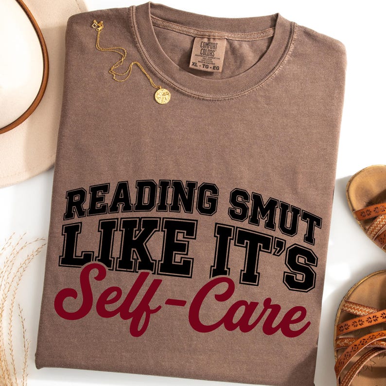 May include: A brown t-shirt with the text "READING SMUT LIKE IT'S Self-Care" in black and burgundy lettering. A gold necklace and a pair of brown sandals are also visible. The shirt has a crew neck and a tag that says "Comfort Colors".