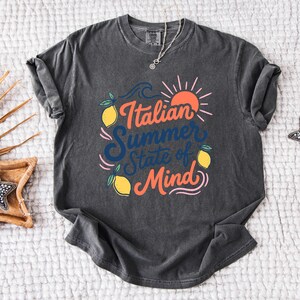 May include: Dark gray t-shirt with the text "Italian Summer State of Mind" in orange and blue. The design includes lemons, a sun, and waves. A silver necklace is visible.