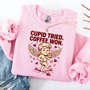 May include: A light pink sweatshirt with the text "CUPID TRIED. COFFEE WON." features a cartoon Cupid holding a coffee mug. The design includes hearts, stars, and an arrow. The sweatshirt is folded on a white surface.