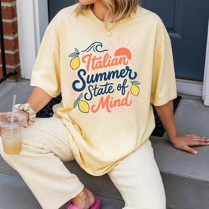 May include: A light yellow t-shirt with the text "Italian Summer State of Mind" in a colorful design featuring lemons, a sun, and waves. The shirt is a relaxed fit and has short sleeves.