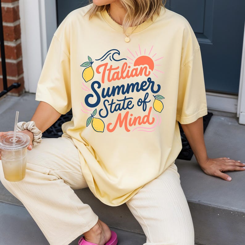 May include: A light yellow t-shirt with the text "Italian Summer State of Mind" in a colorful design featuring lemons, a sun, and waves. The shirt is a relaxed fit and has short sleeves.