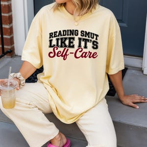 May include: A light yellow t-shirt with the text "READING SMUT LIKE IT'S Self-Care" in black and burgundy lettering. The shirt is a relaxed fit and appears to be made of a soft material. The overall style is casual and comfortable.