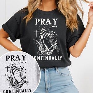 May include: Black t-shirt with the words "PRAY CONTINUALLY" in white. The design features praying hands, a cross, and decorative flourishes. The shirt is a relaxed fit.