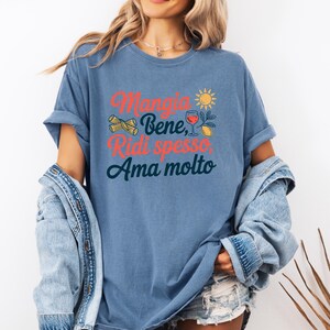 May include: A blue t-shirt with the Italian phrase "Mangia Bene, Ridi spesso, Ama molto" in red and teal lettering. The design includes pasta, a sun, a glass of wine, and a lemon. The shirt has rolled-up sleeves.