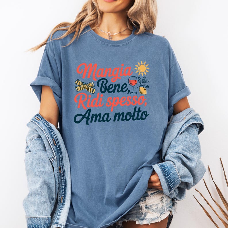 May include: A blue t-shirt with the Italian phrase "Mangia Bene, Ridi spesso, Ama molto" in red and teal lettering. The design includes pasta, a sun, a glass of wine, and a lemon. The shirt has rolled-up sleeves.