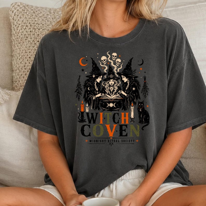 Puede incluir: Una camiseta gris carb&oacute;n con un estampado gr&aacute;fico de tres brujas reunidas alrededor de un caldero. El dise&ntilde;o incluye calaveras, velas, un gato negro y lunas crecientes. El texto dice WITCH COVEN y MIDNIGHT RITUAL SOCIETY en naranja y verde.