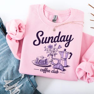 May include: A light pink sweatshirt with the word "Sunday" in dark purple script. Below the word is a graphic of pancakes, a coffee cup, a croissant, a vase of flowers, and a coffee pot. The words "coffee club" are below the graphic.