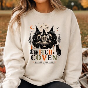 Puede incluir: Una sudadera de cuello redondo color crema con un estampado gr&aacute;fico de tres brujas reunidas alrededor de un caldero. El dise&ntilde;o incluye calaveras, estrellas, lunas, velas y un gato negro, con el texto "WITCH COVEN" y "MIDNIGHT RITUAL SOCIETY".
