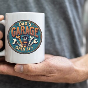 Może przedstawiać: Biały kubek ceramiczny z niebiesko-pomarańczową grafiką, na kt&oacute;rej widnieje napis "Dad's Garage Open 24/7". Grafika przedstawia klucz, śrubę i zębatkę.