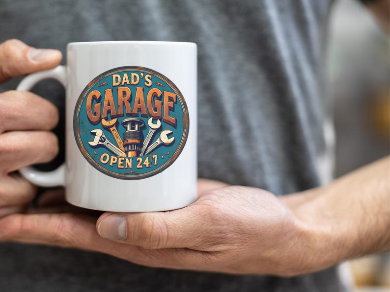 Może przedstawiać: Biały kubek ceramiczny z niebiesko-pomarańczową grafiką, na kt&oacute;rej widnieje napis "Dad's Garage Open 24/7". Grafika przedstawia klucz, śrubę i zębatkę.