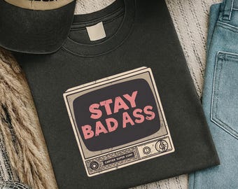 Może przedstawiać: Ciemnoszara koszulka z grafiką w stylu retro telewizora z różowym napisem "STAY BAD ASS". Koszulka jest połączona z czarno-beżową czapką bejsbolową i jasnoniebieskimi dżinsami. Na grafice widnieje napis "CARTIER SUPER CORP."