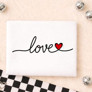 Puede incluir: Un lienzo blanco con la palabra "love" escrita en cursiva negra. La letra "o" es reemplazada por un corazón rojo. Bolas de discoteca plateadas decoran la imagen. El lienzo está sobre una superficie beige.