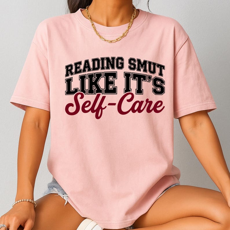 May include: A light pink t-shirt with the text "READING SMUT LIKE IT'S Self-Care" in black and burgundy lettering. The shirt has short sleeves and a relaxed fit, suitable for casual wear.