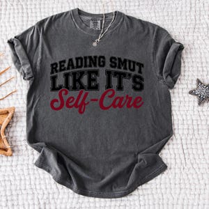 May include: Dark grey t-shirt with the text "READING SMUT LIKE IT'S Self-Care" in black and red lettering. The shirt has a crew neck and short sleeves. A silver necklace and a star-shaped decoration are also visible.
