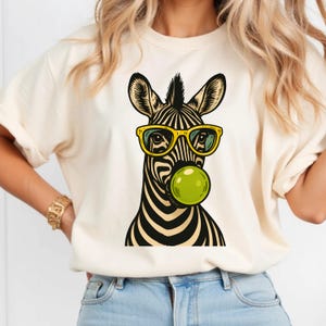 Op de afbeelding: Crèmekleurig T-shirt met een zwart-wit zebra-afbeelding met een gele zonnebril en een groene bubbel. Het zebra-ontwerp heeft een moderne, cartoonachtige stijl. Het shirt is gemaakt van een zacht materiaal.