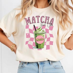 May include: Cream-colored t-shirt featuring a graphic of a matcha iced latte. The design includes the word "Matcha" in pink, a green drink with whipped cream, and a pink and white checkered background.