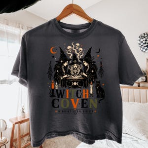 Puede incluir: Una camiseta gris carb&oacute;n con un estampado gr&aacute;fico de tres brujas con sombreros puntiagudos reunidas alrededor de un caldero y un libro abierto. El dise&ntilde;o incluye calaveras, velas, un gato negro y el texto WITCH COVEN MIDNIGHT RITUAL SOCIETY.
