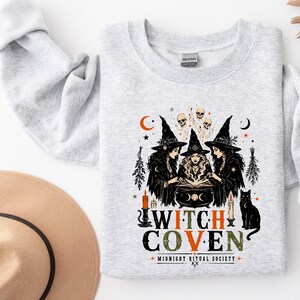 Puede incluir: Una sudadera gris claro de cuello redondo con un estampado gr&aacute;fico de tres brujas alrededor de un caldero, calaveras, velas y un gato negro. El texto "WITCH COVEN" aparece en letras grandes con "MIDNIGHT RITUAL SOCIETY" impreso debajo en negro y naranja.