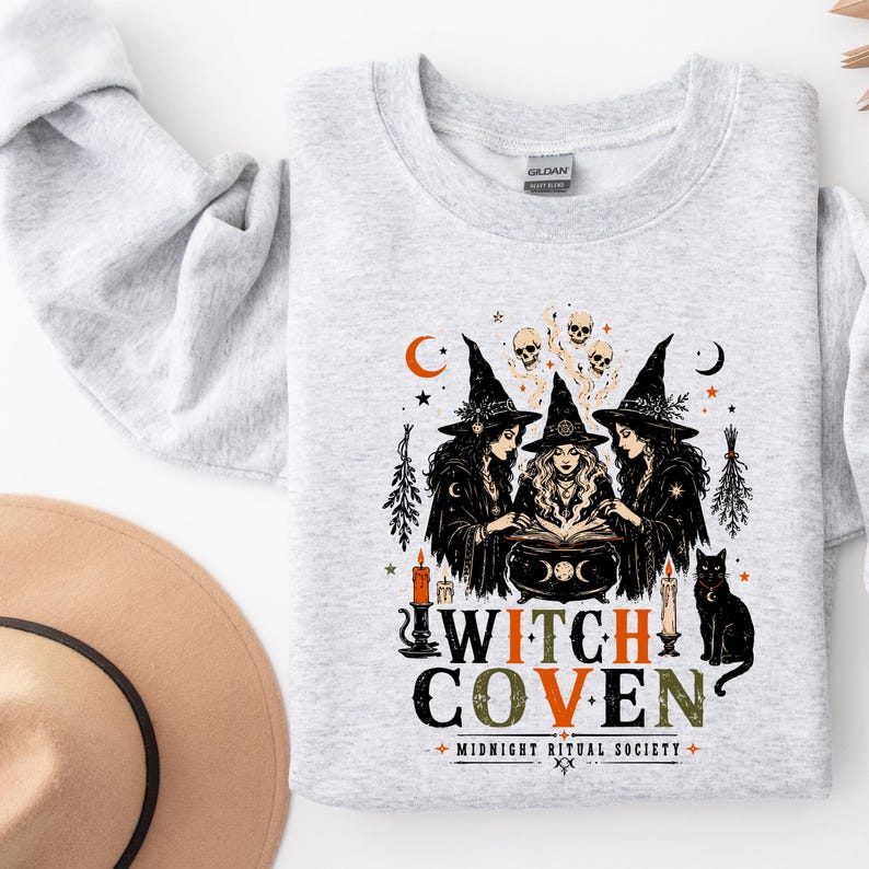 Puede incluir: Una sudadera gris claro de cuello redondo con un estampado gr&aacute;fico de tres brujas alrededor de un caldero, calaveras, velas y un gato negro. El texto "WITCH COVEN" aparece en letras grandes con "MIDNIGHT RITUAL SOCIETY" impreso debajo en negro y naranja.
