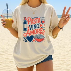 May include: Cream-colored t-shirt with the words "Pasta Vino Amore" in red and blue. The design includes a wine bottle, a glass of wine, a heart, and a bowl of pasta. The shirt is worn on a beach.
