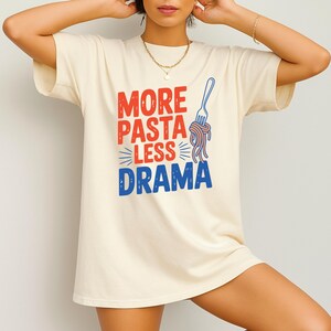 May include: Cream-colored t-shirt with the phrase "MORE PASTA LESS DRAMA" in red and blue, accompanied by a blue fork and spaghetti graphic. The shirt is a casual, comfortable style.