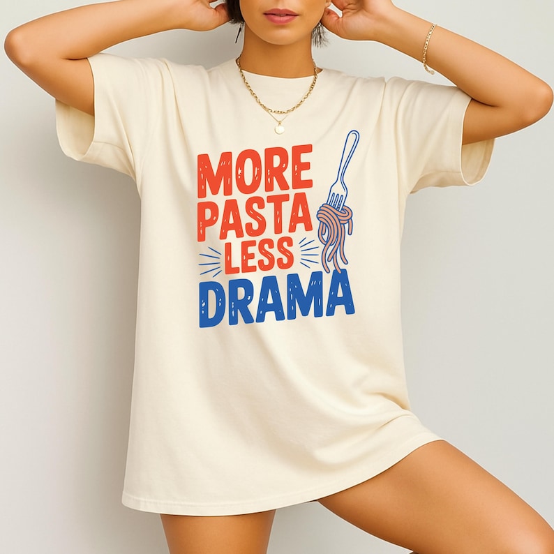 May include: Cream-colored t-shirt with the phrase "MORE PASTA LESS DRAMA" in red and blue, accompanied by a blue fork and spaghetti graphic. The shirt is a casual, comfortable style.