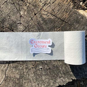 May include: A roll of white toilet paper with a sticker that reads "Excrement Occurs" in red text. The sticker also features a small illustration of a toilet paper roll. The toilet paper is unrolled on a textured surface.