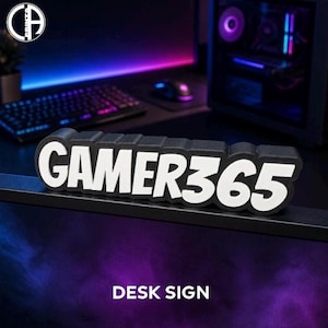 May include: A black desk sign featuring the white text GAMER365 in a bold, sans-serif font. The sign has a thick, raised profile with a matte finish and is positioned horizontally on a flat surface, displaying the text clearly against the dark background.