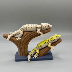 Puede incluir: Dos figuras articuladas de gecko, una de color crema con manchas marrones y otra amarilla con manchas negras, descansan sobre una estructura marr&oacute;n en forma de &aacute;rbol. La base es un rect&aacute;ngulo azul oscuro. Los geckos posan como si estuvieran descansando en una rama.