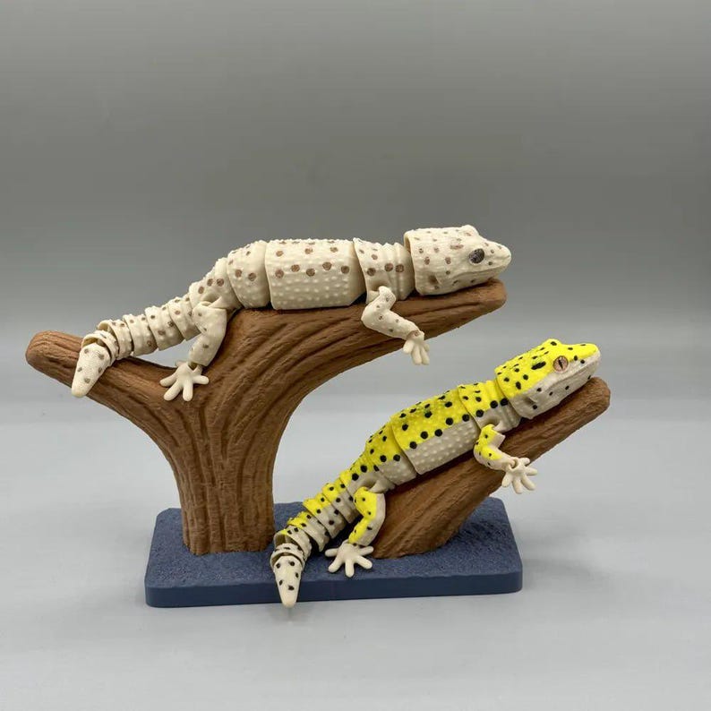 Puede incluir: Dos figuras articuladas de gecko, una de color crema con manchas marrones y otra amarilla con manchas negras, descansan sobre una estructura marr&oacute;n en forma de &aacute;rbol. La base es un rect&aacute;ngulo azul oscuro. Los geckos posan como si estuvieran descansando en una rama.