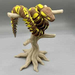 Puede incluir: Un juguete de serpiente articulada marr&oacute;n y amarilla enrollada en un soporte beige con forma de &aacute;rbol. La serpiente tiene una cabeza y una cola detalladas, con segmentos amarillos y marrones. El soporte tiene un aspecto texturizado y naturalista.