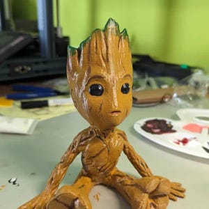 May include: A 3D printed figurine of Groot from Guardians of the Galaxy. The figurine is painted in a light brown color with green accents. Groot is sitting with his legs crossed and his arms resting on his knees.