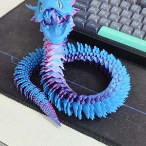 Może przedstawiać: Drukowana w 3D figurka smoka w kolorze niebieskim i fioletowym, z kolczastym, segmentowym ciałem i otwartą paszczą z zębami.