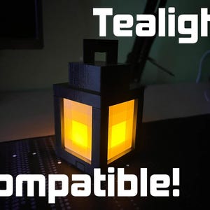 May include: Black, cube-shaped tealight holder with a warm yellow glow emanating from within. The words "Tealight compatible!" are printed in white. The lantern has a handle on top and a pixelated design.