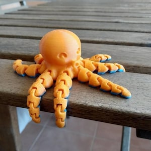 Può includere: Una statuetta di un polpo arancione con braccia segmentate e accenti blu. Il polpo è seduto su una superficie di legno.