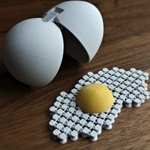 Op de afbeelding: Een witte plastic eier-vormige container met een geel plastic ei erin. De container is open en het gele ei ligt op een wit plastic rooster.