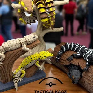 以下が含まれることがあります： 茶色、黄色、黒、白などの様々な色のヘビやヤモリなどの関節式爬虫類フィギュアの展示。 フィギュアは人工の枝の上に配置され、本物のような外観を作り出しています。 下部には「TACTICAL KAOZ」の文字が見えます。