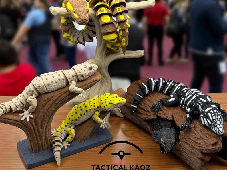 Puede incluir: Exhibici&oacute;n de figuras articuladas de reptiles, incluyendo serpientes y geckos, en varios colores como marr&oacute;n, amarillo, negro y blanco. Las figuras est&aacute;n posadas sobre ramas artificiales, creando una apariencia realista. El texto "TACTICAL KAOZ" es visible en la parte inferior.