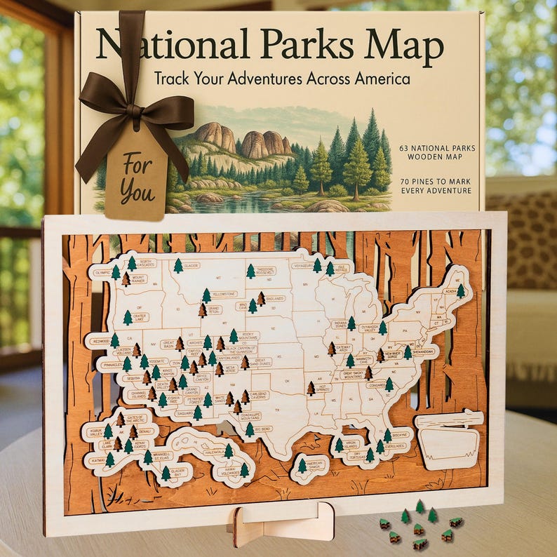 Puede incluir: Un mapa de madera de los Parques Nacionales de Estados Unidos, con 63 parques y 70 marcadores de pinos. El mapa est&aacute; enmarcado en madera, con una caja que muestra "National Parks Map" y "Track Your Adventures Across America". Una cinta marr&oacute;n y una etiqueta "For You" adornan la caja.