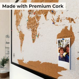 May include: A framed world map made of cork with the text Made with Premium Cork. The map features brown landmasses on a light background, accented with colorful push pins and a vertical photograph of two people pinned over the Australia region.