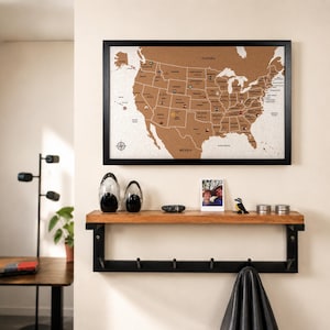 May include: A rectangular cork map of the United States in a black frame, mounted above a wooden shelf with a black metal base and six hooks. The map features state outlines, labels, and small circular markers placed across various regions.