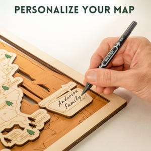 Puede incluir: Mapa de madera con contornos de estados y un marco. El texto "PERSONALIZA TU MAPA" est&aacute; en la parte superior. Una mano usa un marcador negro para escribir en un letrero de madera que dice "Anderson Family".
