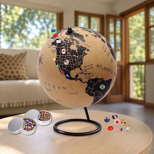 May include: A cork globe featuring a black printed world map, mounted on a black metal stand. The globe displays various national flag pins and colorful push pins. Two small silver metal tins containing additional push pins are placed nearby.