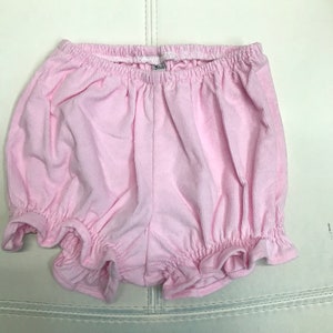 May include: Pink baby bloomers with a ruffled hem. The bloomers are made of a soft, ribbed fabric.
