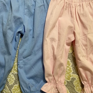 May include: A pair of light blue pants with an elastic waistband and a pair of light pink pants with an elastic waistband and ruffled cuffs. Both garments feature a simple design with gathered fabric at the waist and are shown side by side.