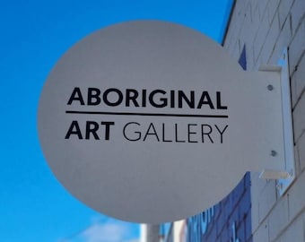 May include: A white, circular sign for an Aboriginal Art Gallery is mounted on a brick wall. The sign has black text that reads "ABORIGINAL ART GALLERY". The sky is blue in the background.