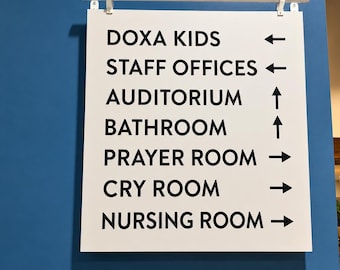 May include: A white rectangular directional sign with black text and arrows mounted on a blue wall. The sign lists: DOXA KIDS, STAFF OFFICES, AUDITORIUM, BATHROOM, PRAYER ROOM, CRY ROOM, and NURSING ROOM. A small blue sign below reads DOXA WELCOME.