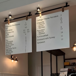 Könnte beinhalten: Zwei weiße, rechteckige Menütafeln, die an schwarzen Metallstangen mit beigen Lederriemen hängen. Die Tafeln zeigen schwarzen Text mit einer Liste verschiedener Kaffeespezialitäten, Tees und Speisen wie Pfannkuchen, Omeletts und Sandwiches mit Preisen.